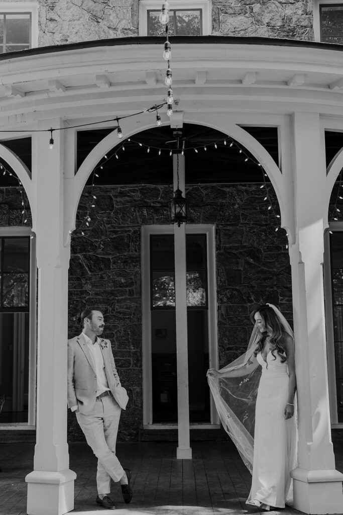 A bride and groom standing on a victorian porch. Both are leaning against a post. The groom is leaning with his legs crossed, hands in his pockets, and looking at the bride. The bride is leaning while holding her veil