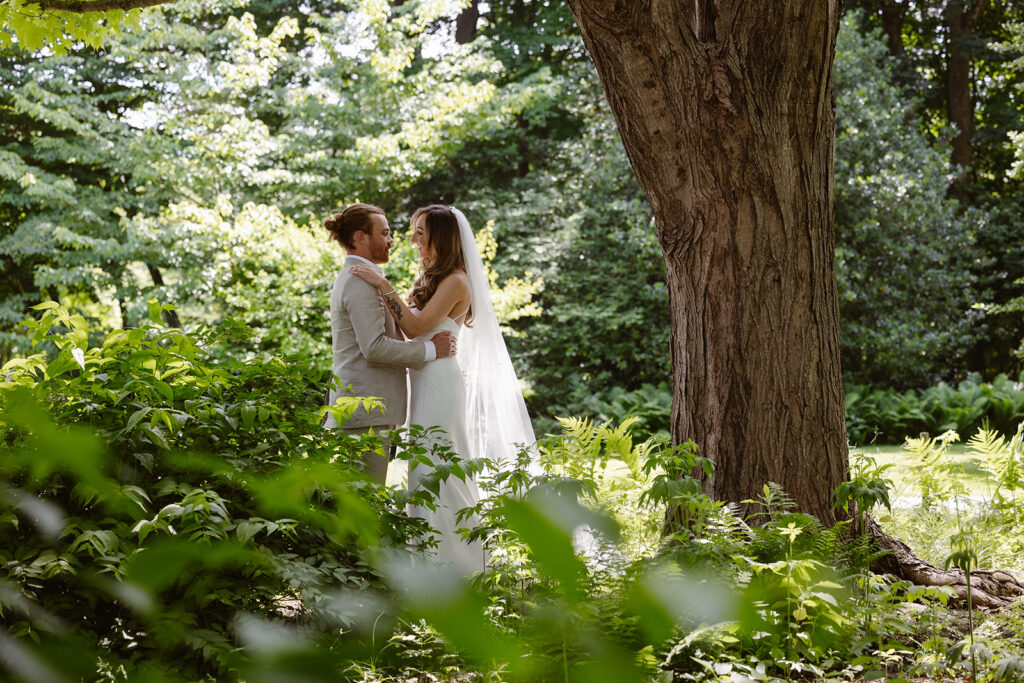 A bride and groom on their wedding day standing under an old tree surrounded by lush greens. The couple is facing each other, and looking at each other lovingly