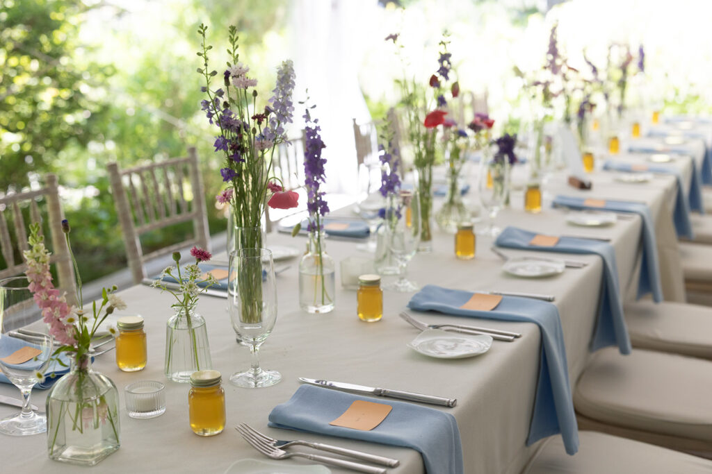 A photo of the table scape from the reception of a bride and groom. There are pink and purple wildflowers in glass vases, jars of honey at every seat, and light blue napkins with a peach name card