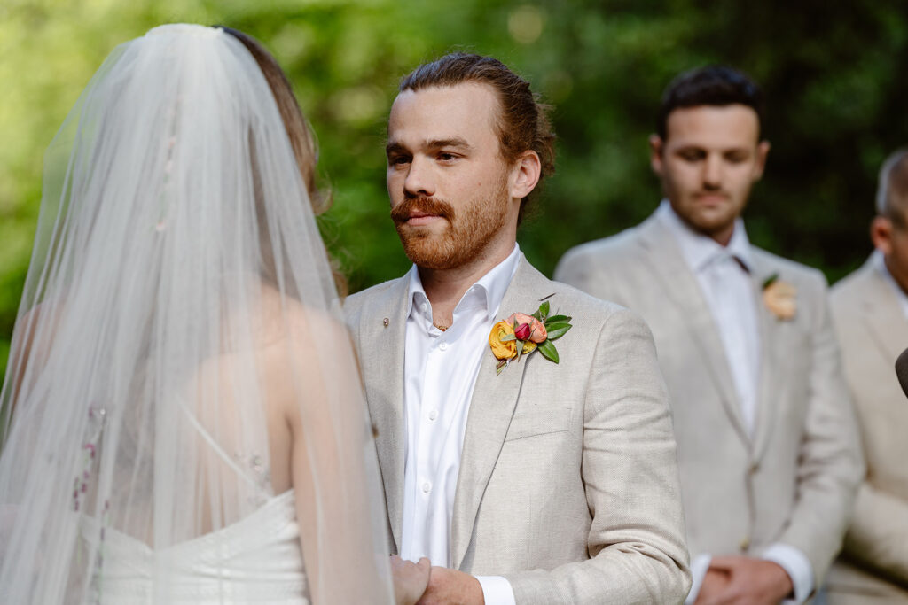 A groom listening to his bride say her vows during their wedding ceremony. The bride is wearing a Boho dress with a beautiful veil. The groom is wearing a tan linen suit with a brightly colored boutonniere