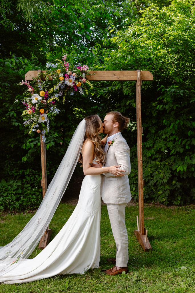 A bride and groom sharing their first kiss as a married couple during their wedding ceremony.  The bride is wearing a Boho dress with a long custom veil, and the groom is wearing a tan linen suit. They are standing under a hand made arbor adorned with gorgeous summer blooms