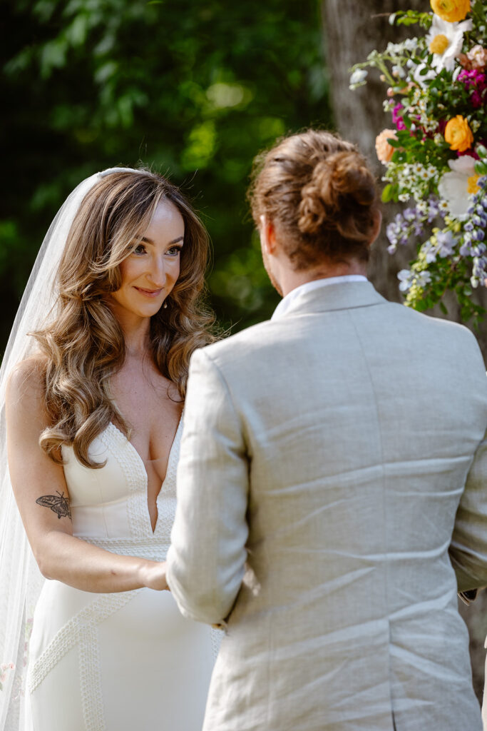 A bride listening to her groom say his vows during their wedding ceremony. The bride is wearing a Boho dress with a beautiful veil. The groom is wearing a tan linen suit