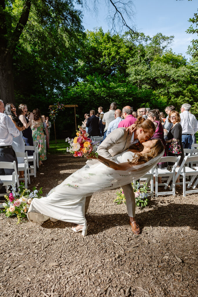 A bride and groom take a moment at the end of the aisle to dip  and kiss to celebrate saying I do. The bride is wearing a Boho dress with a long custom veil, and the groom is wearing a tan linen suit.  The bride is holding the most beautiful summer bouquet