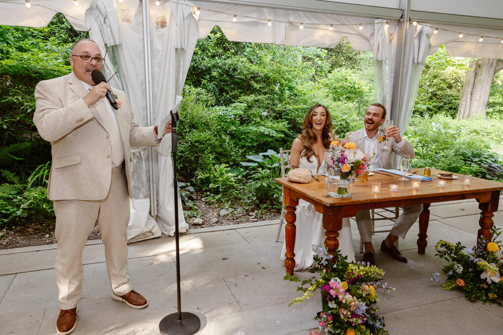 A bride and groom laughing during a speech at their reception. They are sitting at a wooden table under a tent. The dad is giving a speech to the couple. He is standing at a microphone