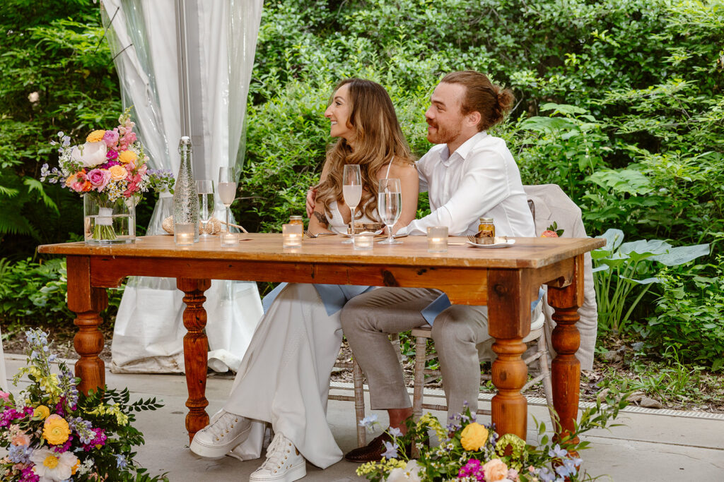 A bride and groom smiling during a speech at their reception. They are sitting at a wooden table under a tent. There are beautiful flower arrangements at their feet. The bride is wearing a gorgeous Boho dress, and is wearing white sneakers. The groom has his arm around the bride