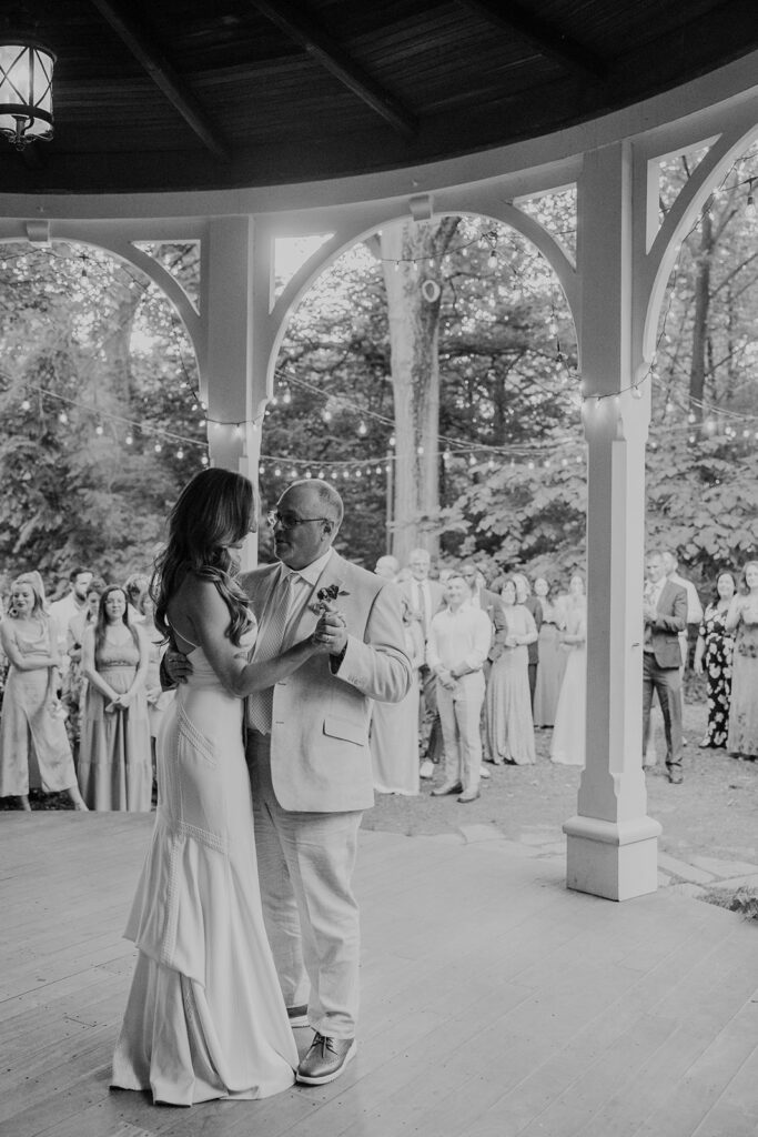 A bride dancing with her father on a victorian wraparound porch. The photo is in black and white. There are twinkly lights, and the attendees of the wedding are in the background watching the dance