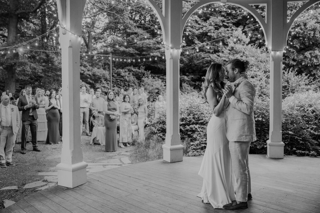 A black and white photo of the bride and groom during their first dance  on their wedding day. They are dancing on a victorian porch surrounded by loved ones and twinkly lights. 