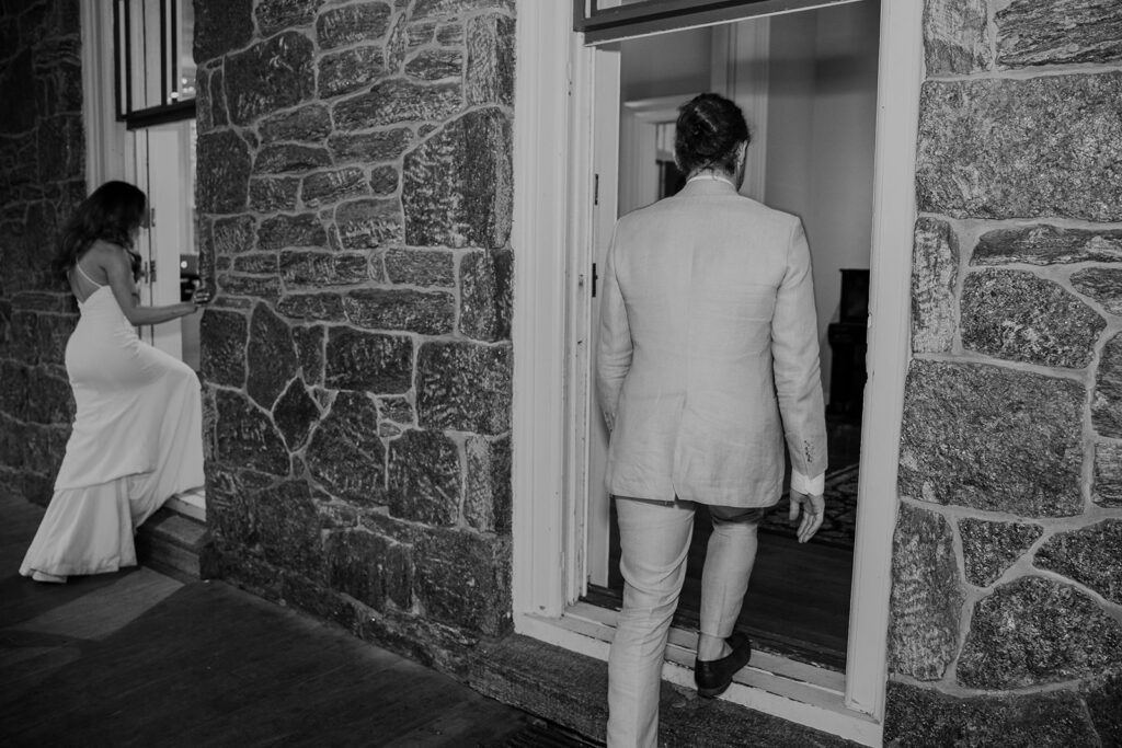 A black and white photo of the bride and groom walking into opened floor to ceiling windows of a victorian mansion. The couple are walking in the windows to get the party started during their reception