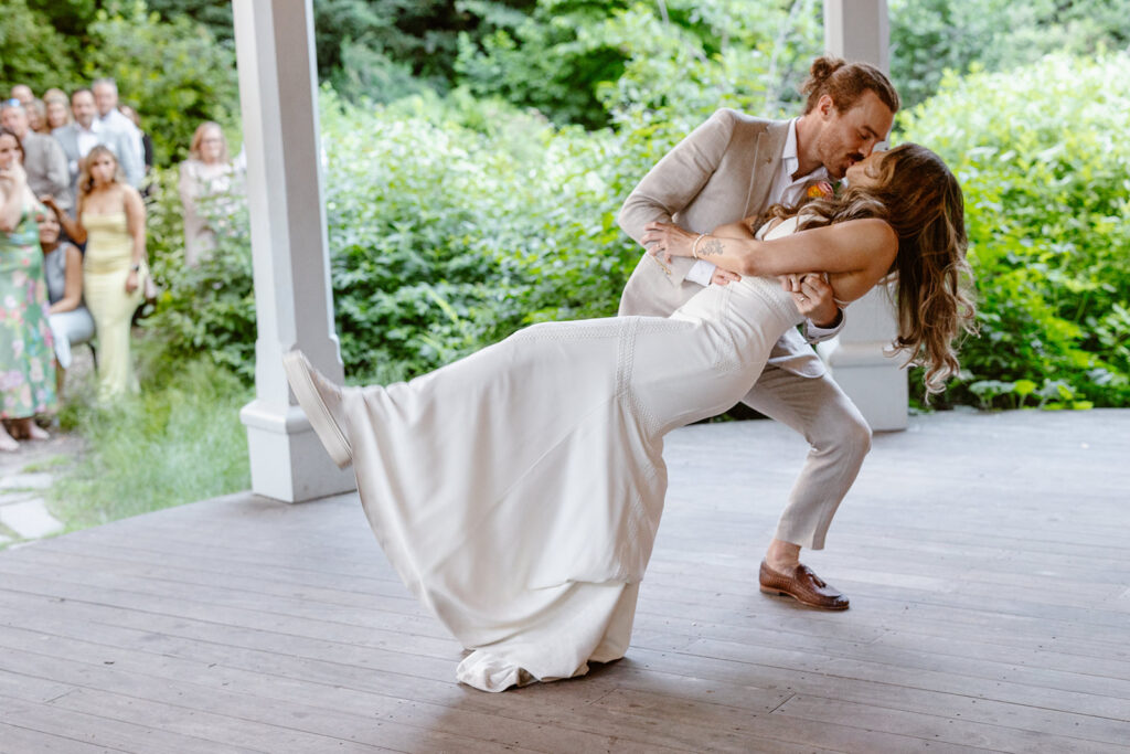 A bride and groom doing a dip-kiss at the end of their first dance on their wedding day. The bride is wearing a Boho dress and sneakers, and the groom is wearing a tan linen suit with leather loafers. 