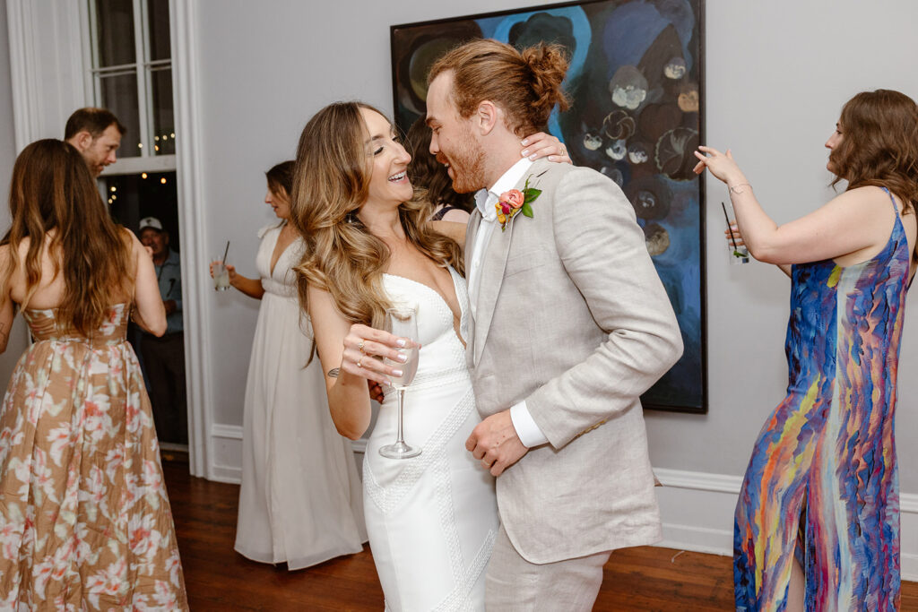 Bride and groom dancing during their reception. The bride is holding aq glass of wine wile they dance. They are in a room with paintings on the wall