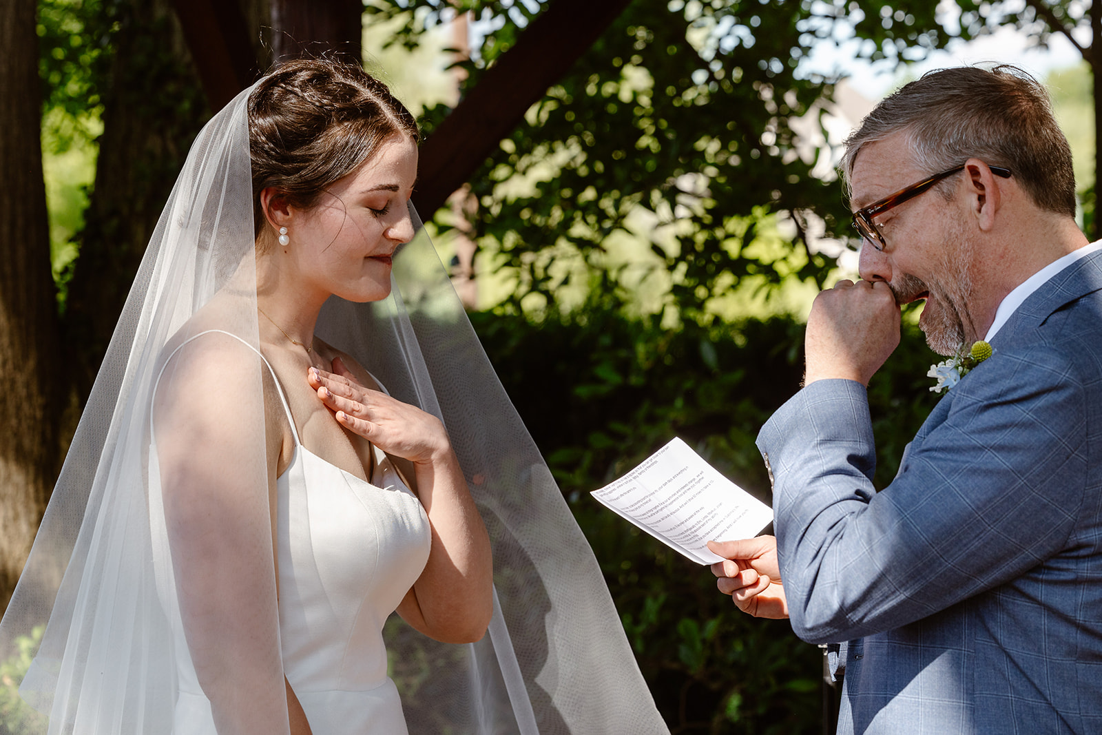 A bride and groom during their vows. The groom is reading his vows, and the brides eyes are closed with emotion. She is holding her hand to her heart.