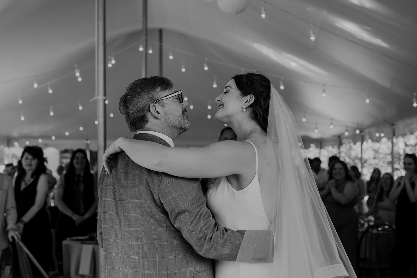 A black and white photo of a bride and groom having their first dance at their wedding/ They are under a tent, and their are twinkly lights