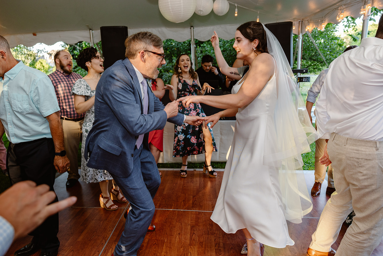 A couple dancing at their wedding reception the woman is wearing a white wedding dress and veil and is dancing with her husband who is wearing a blue suit and behind them their guests are also dancing