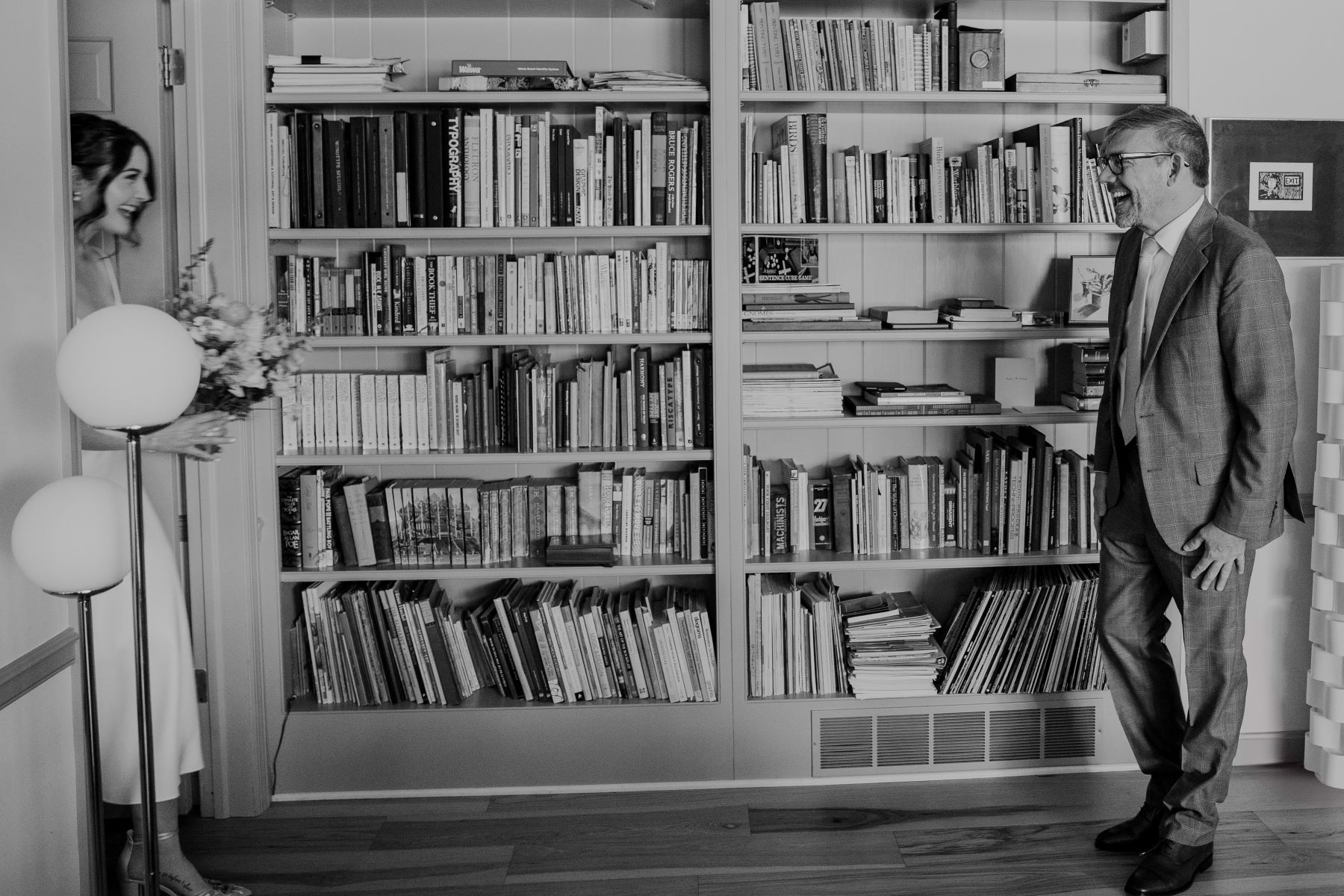 A couple having a first look in their home library the man is smiling as the woman is a white wedding dress holding a bouquet of flowers walks through the door 