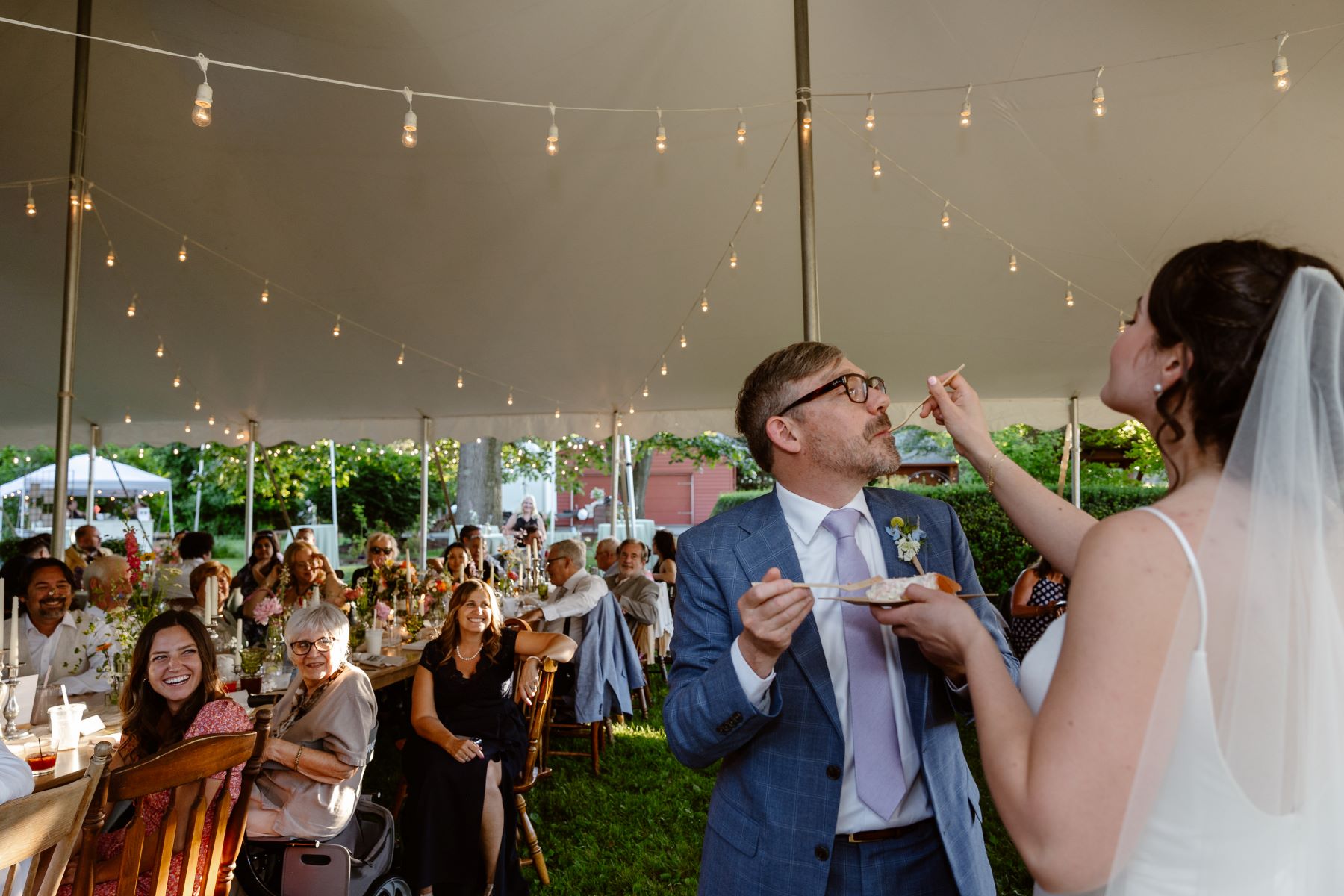 A couple at their wedding reception their guests are all seated at a long wooden table and they are having their ceremony in a white tent the woman is wearing a white dress and veil and feeding cake to her partner who is wearing a blue suit and purple tie 