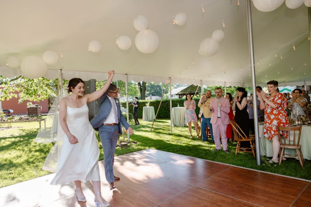 A couple celebrating their marriage and walking onto a wooden dance floor together the woman is wearing a white dress and veil and the man is wearing a blue suit and they are holding one arm each up in the air while their guests are cheering 