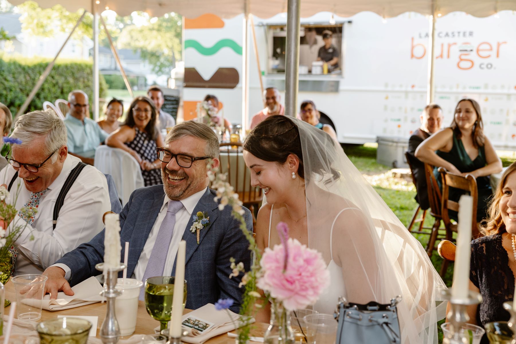 A couple sitting at a wedding table laughing together the woman is wearing a white wedding dress and veil and the man is wearing a blue suit and they are surrounded by their wedding guests