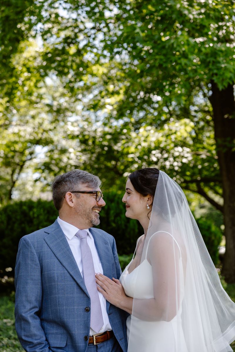 A couple smiling at each other the woman is wearing a white wedding dress and veil and is touching her partner's chest the man is wearing a blue suit and glasses and has his hands in his pockets