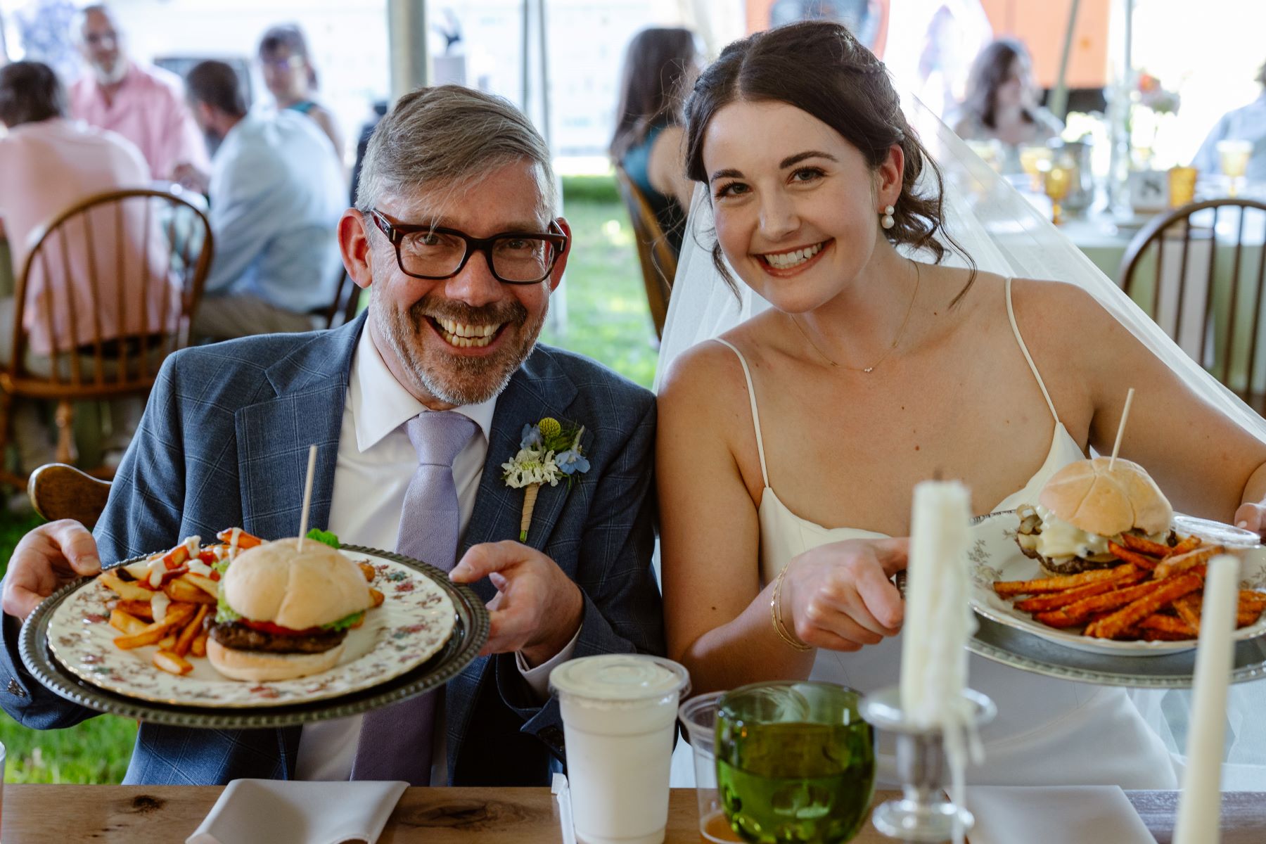 A couple at their wedding dinner showing their plates of food the woman is wearing a white dress and white veil and the man is wearing a blue suit and glasses they are both smiling 