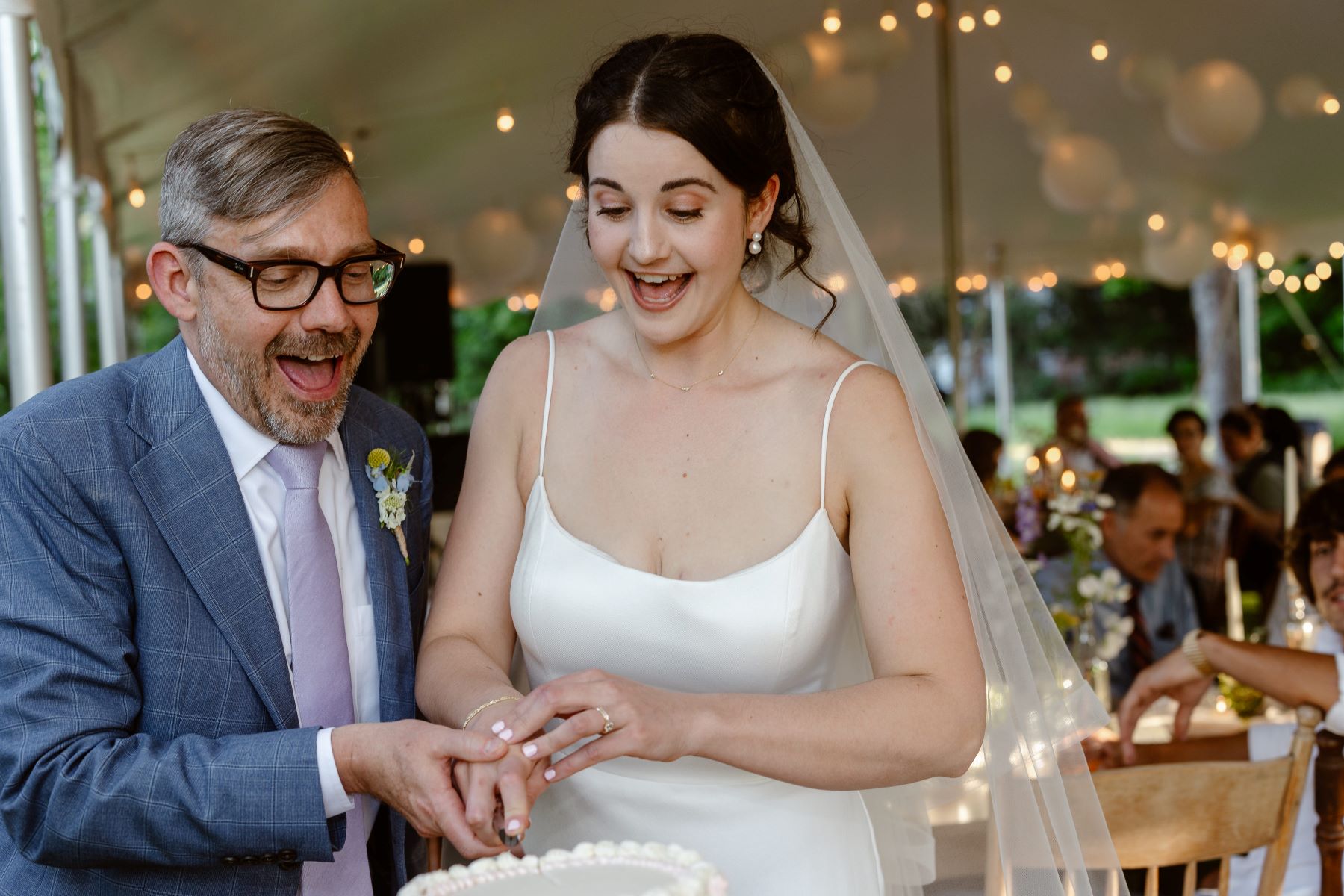 A couple cutting their wedding cake and smiling the woman is wearing a white veil and white wedding dress the man is wearing glasses and a blue suit 