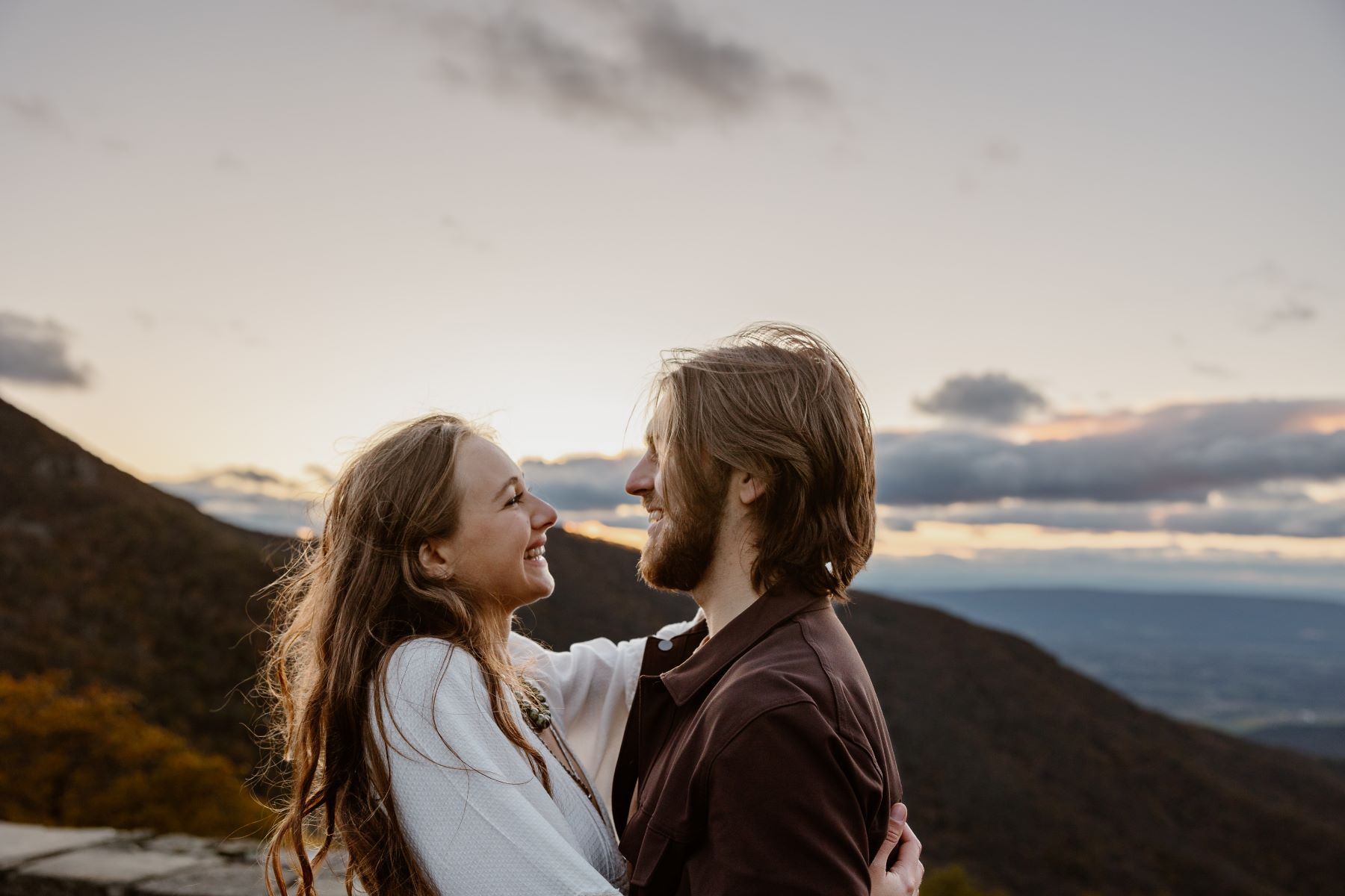 A couple smiling at each other and laughing the woman is wearing a white shirt and the man is wearing a brown shirt and behind them are the mountains and a valley