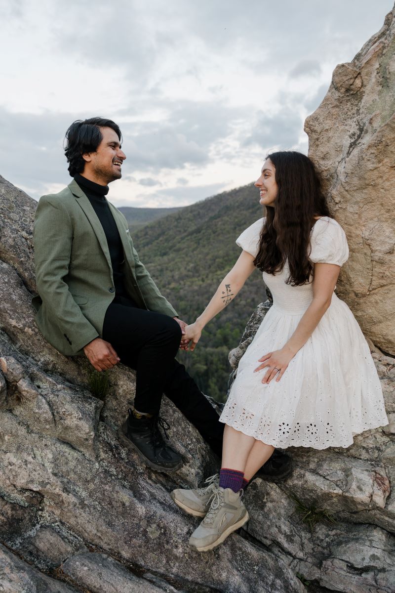 A couple holding hands and sitting on a rock and smiling at each other the woman is wearing a white dress and sneakers and the man is wearing all black with a green jacket top 