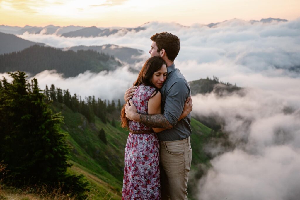 A couple embraces at the top of a mountain during their elopement at sunrise. The couple is above the clouds.