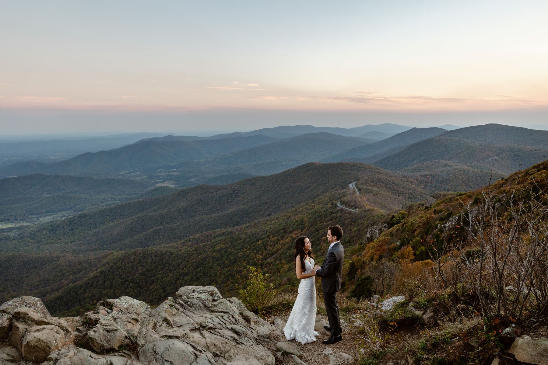 A couple standing on rocks overlooking the mountains the woman is wearing a white wedding dress and holding her partner's hands and he is wearing a black tuxedo 