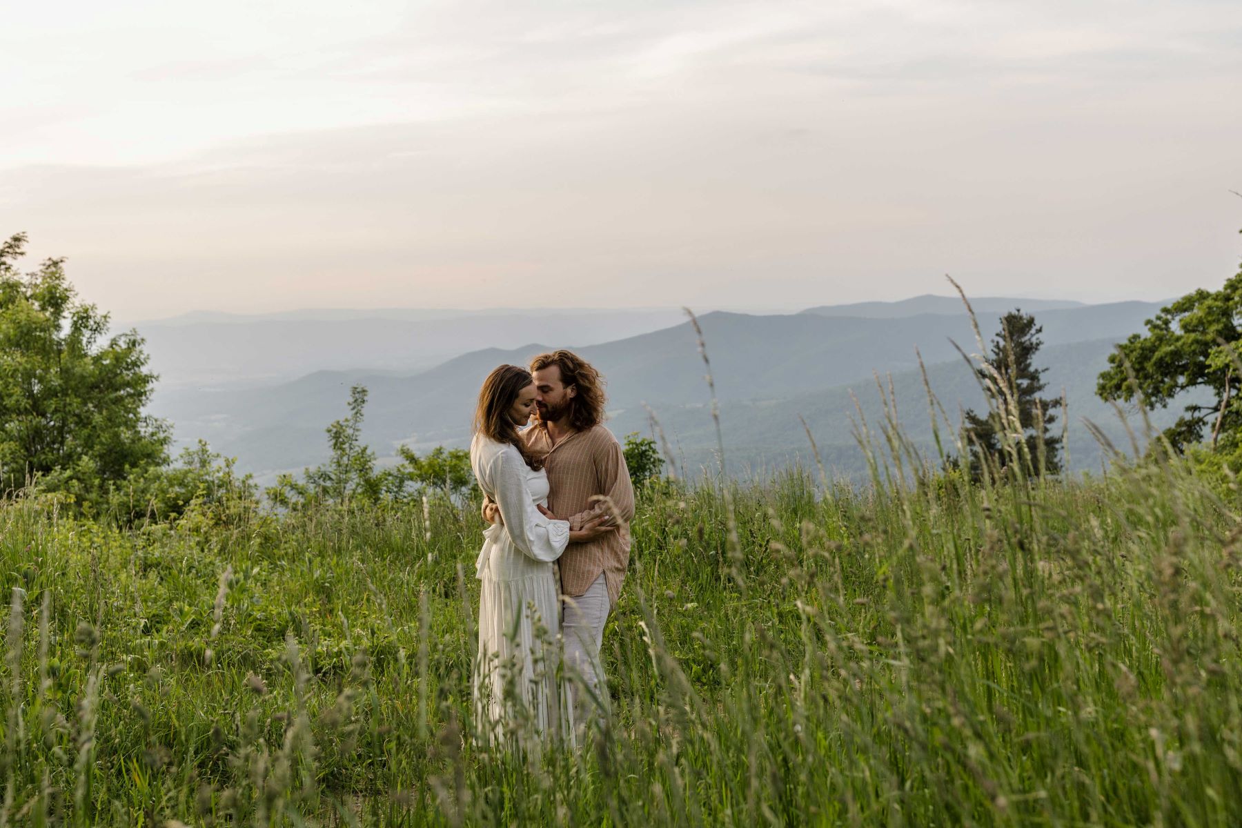 A couple hugging each other in a field of grass and behind them are the mountains the woman is wearing a white dress top and a white skirt and her partner is wearing a brown loose shirt and brown pants 