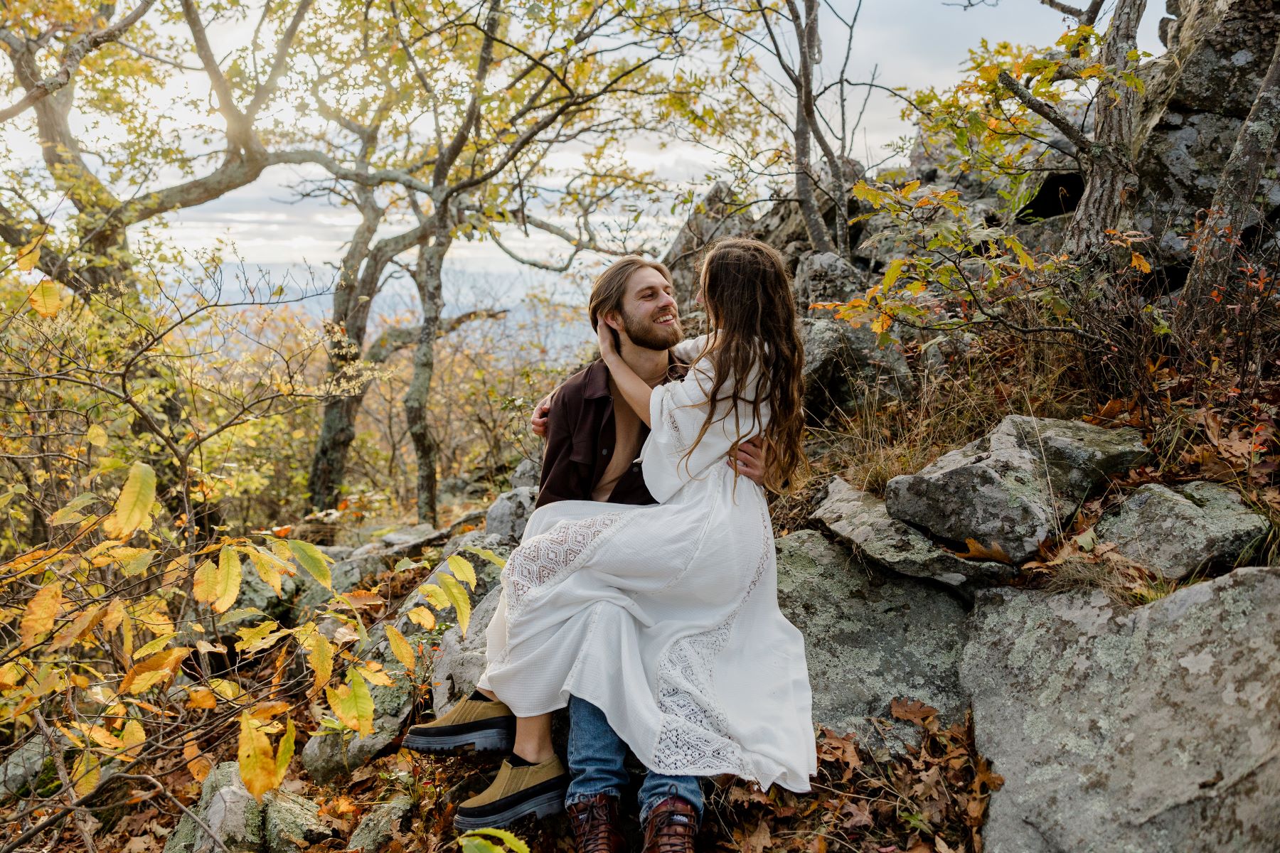 A man is sitting against a rock and his partner who is wearing a white dress is sitting on his lap and both of them are looking at each other and smiling and they are surrounded by fall foliage