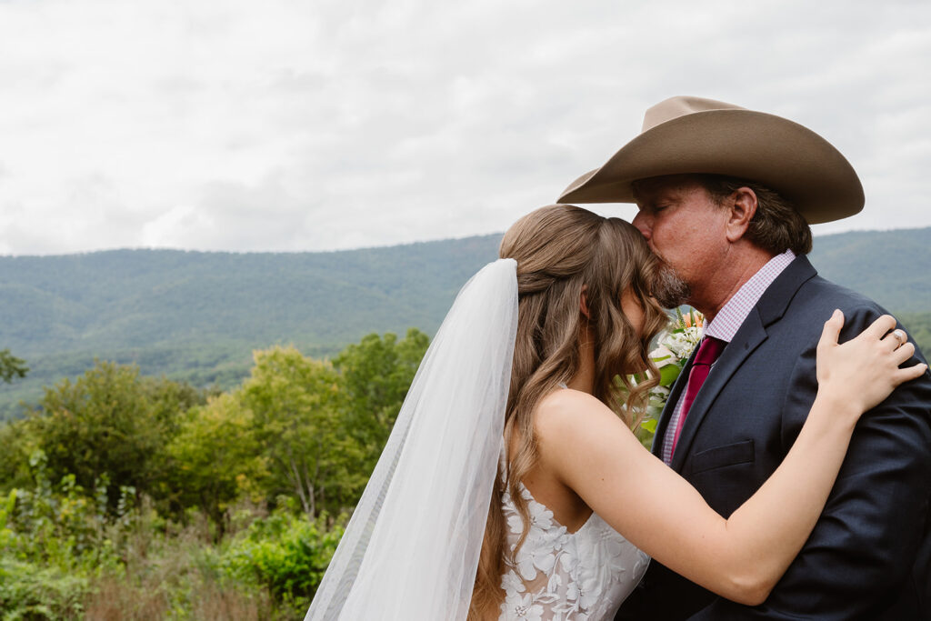 A dad sees his daughter for the first time during her wedding day. He is wearing a cowboy hat, and is kissing his daughters forehead. She os wearing a dress with lace, and a long veil