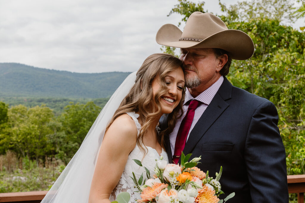 A dad sees his daughter for the first time during her wedding day. He is wearing a cowboy hat, and is kissing his daughters forehead. She os wearing a dress with lace, and a long veil