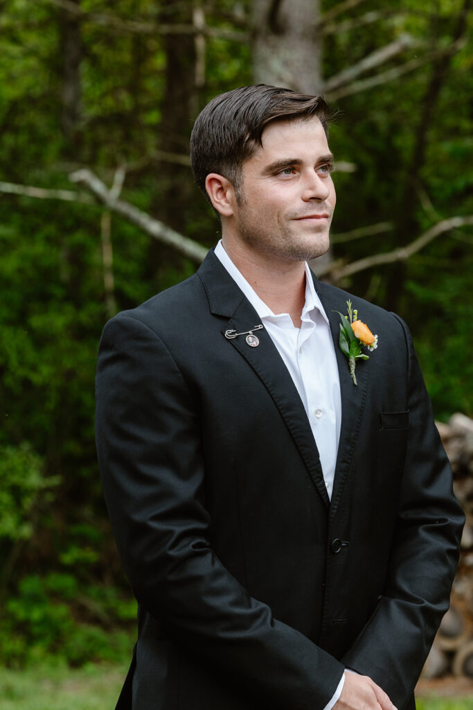 A groom watching his bride walk down the aisle towards him. He has a slight smile on his face. He is wearing a black jacket, white shirt, and a sentimental pin 