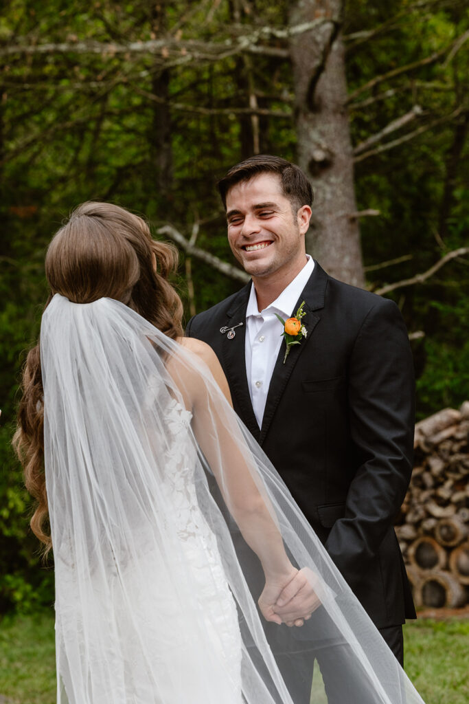 A bride and groom hold hands during their intimate wedding ceremony. She is wearing a dress with lace and a long veil, and he is wearing a black suit with an orange flower. The groom is laughing