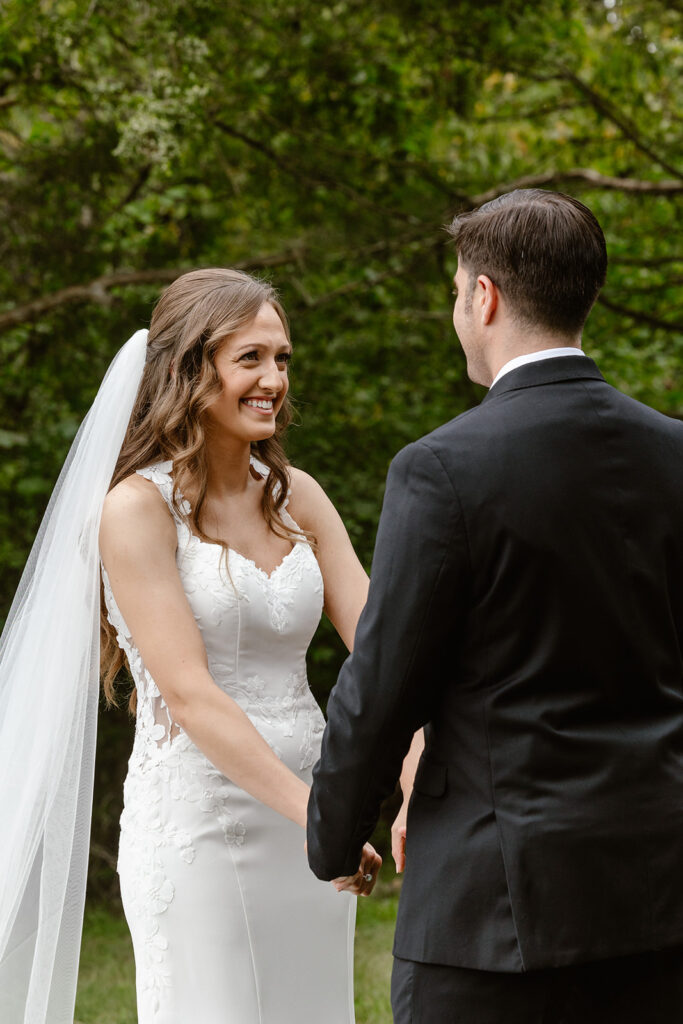 A bride and groom hold hands during their intimate wedding ceremony. She is wearing a dress with lace and a long veil, and he is wearing a black suit with an orange flower. The bride is smiling ear to ear