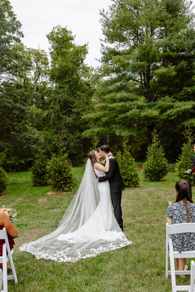 A bride and groom during the first kiss of their wedding ceremony. The bride is wearing a long lace veil, and the groom is wearing a black suit