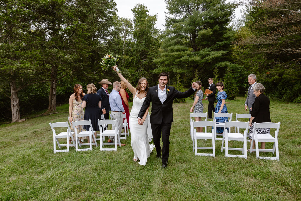 A bride and groom walk down the aisle with their arms in the air with excitement after eloping in front of their immediate families