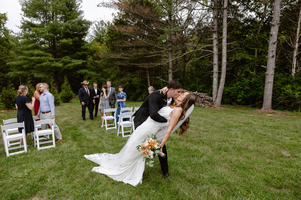 The bride and groom dip kiss at the end of the aisle after being announced. There are 9 guests watching.