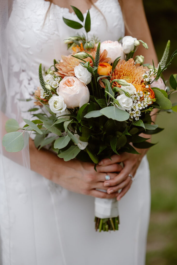 A closeup photo of a bride holding her bouquet. The bouquet is made up of white and peachy colored flowers