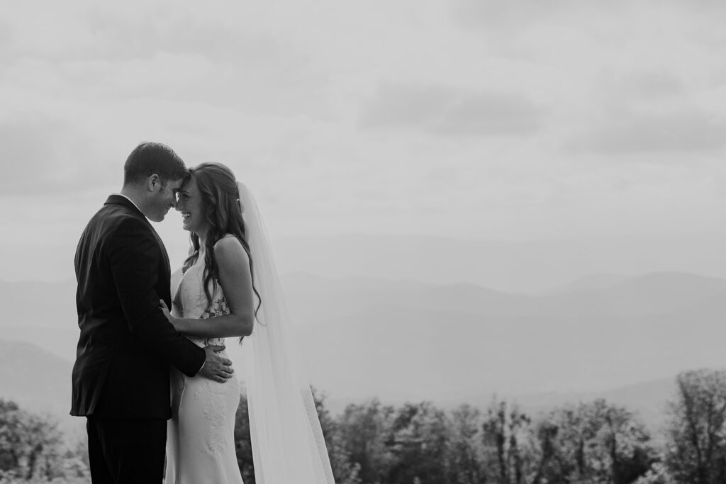 A black and white photo of a bride and groom face each other with their foreheads touching. You can see the mountains in the background. The couple is beaming