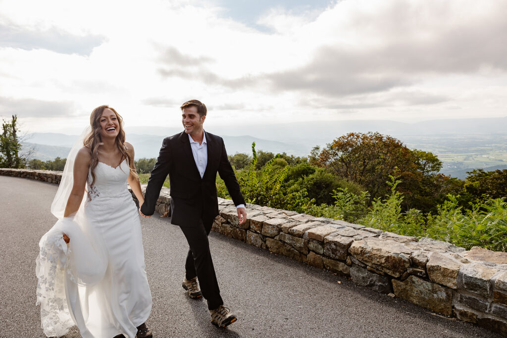 A bride and groom walking hand in hand on Skyline Drive. The bride is holding up her long lace veil. Both the bride and groom are laughing