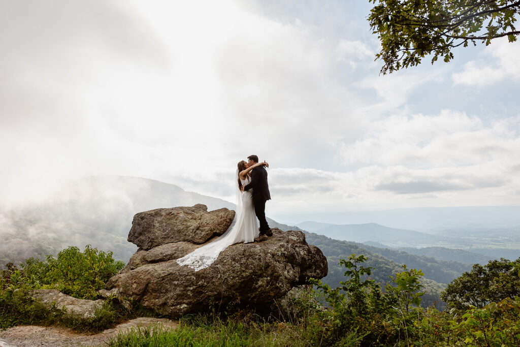 A couple embraces and kisses at an overlook along Skyline Drive in Shenandoah National Park. They are standing on a giant rock formation, and you can see the mountains in the distance