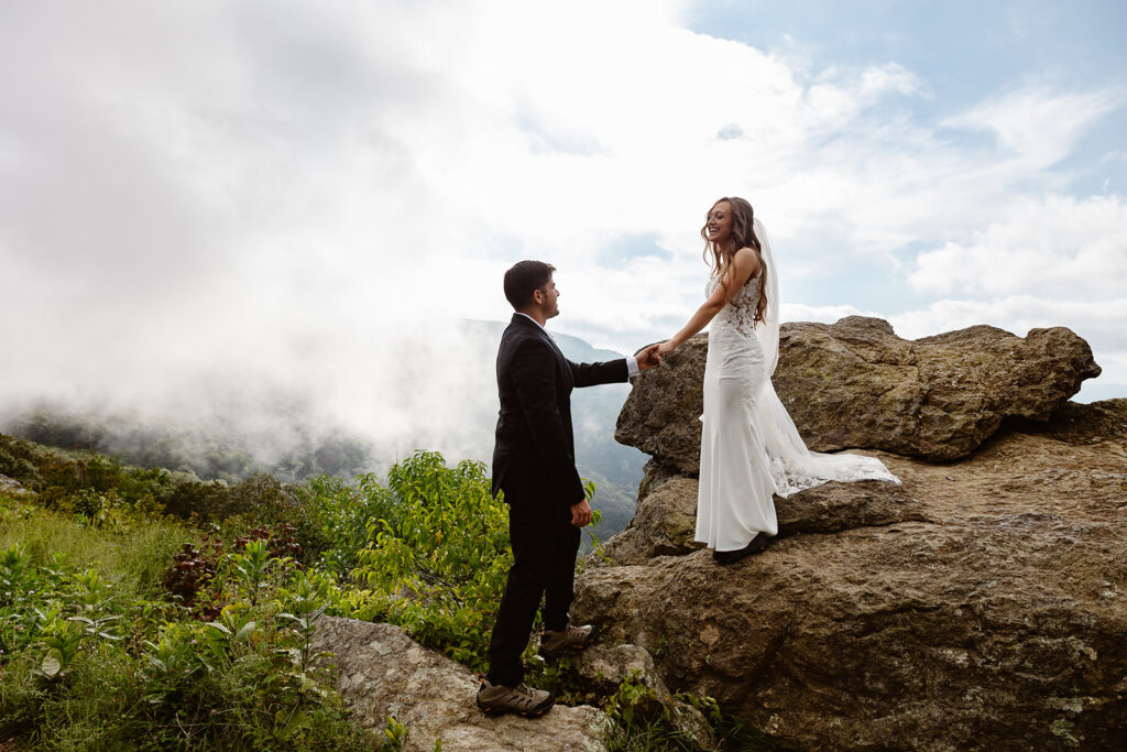 A groom helps his bride down from a large rock. The bride is smiling, She is wearing a lace veil, and he is wearing a black suit