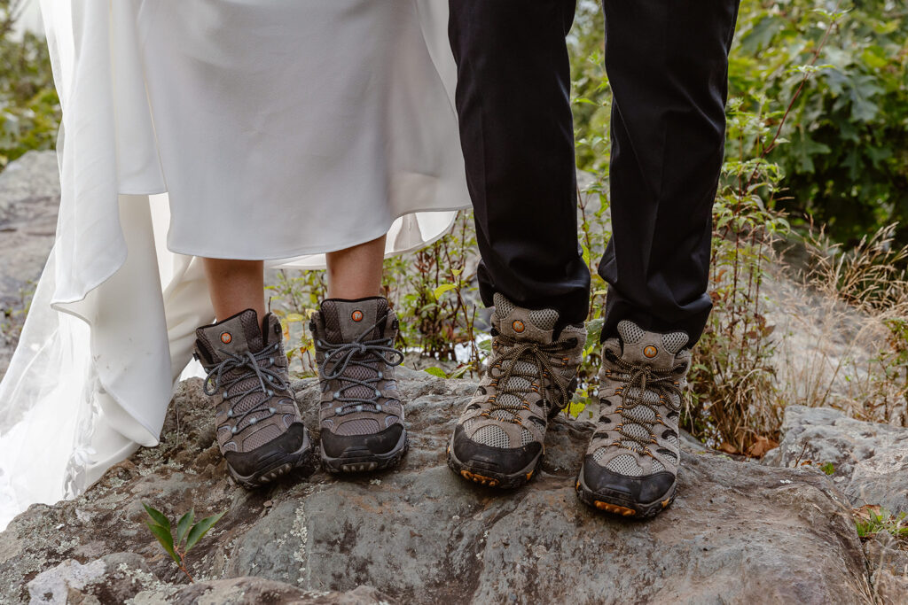 A closeup of a bride and grooms hiking boots. The couple is standing on rock, and the bride is holding up her dress so you can see her boots