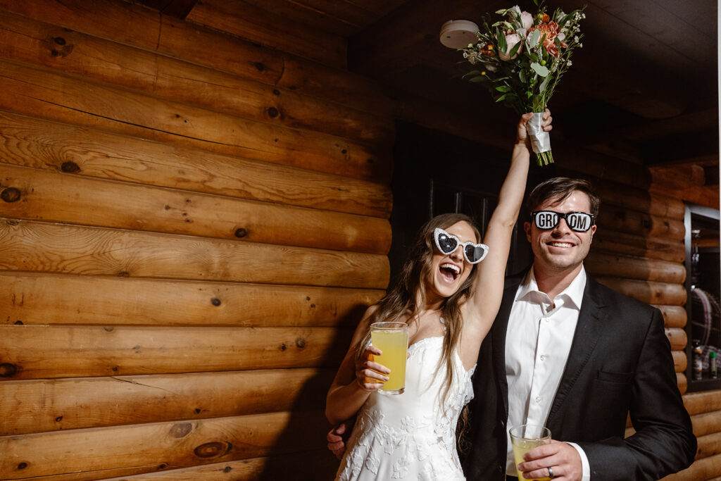 A bride and groom end their night with drinks and fun sunglasses. The bride is holding her bouquet in the air and cheering