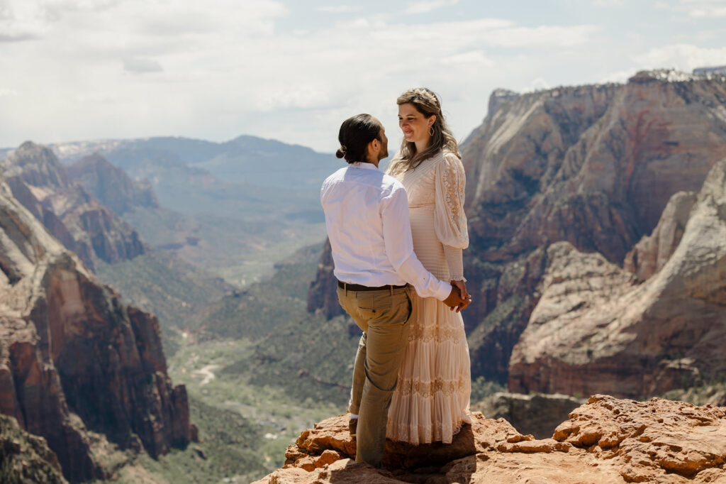 A couple standing on top of a cliff at Observation Point in Zion National Park. They are holding hands and smiling at each other the woman is wearing a dress and the man is wearing awhite button up and tan pants