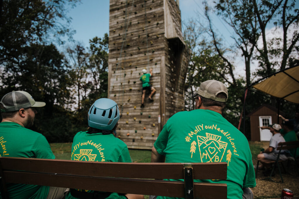 Three people in the foreground watch someone on on a rock climbing wall at a summer camp. The three people are wearing matching green shirts that were given to all of the guests of the wedding