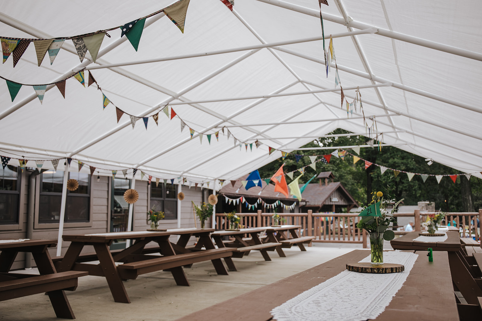 A large white tent with colorful flags hanging from the ceiling and large wooden picnic tables and benches 