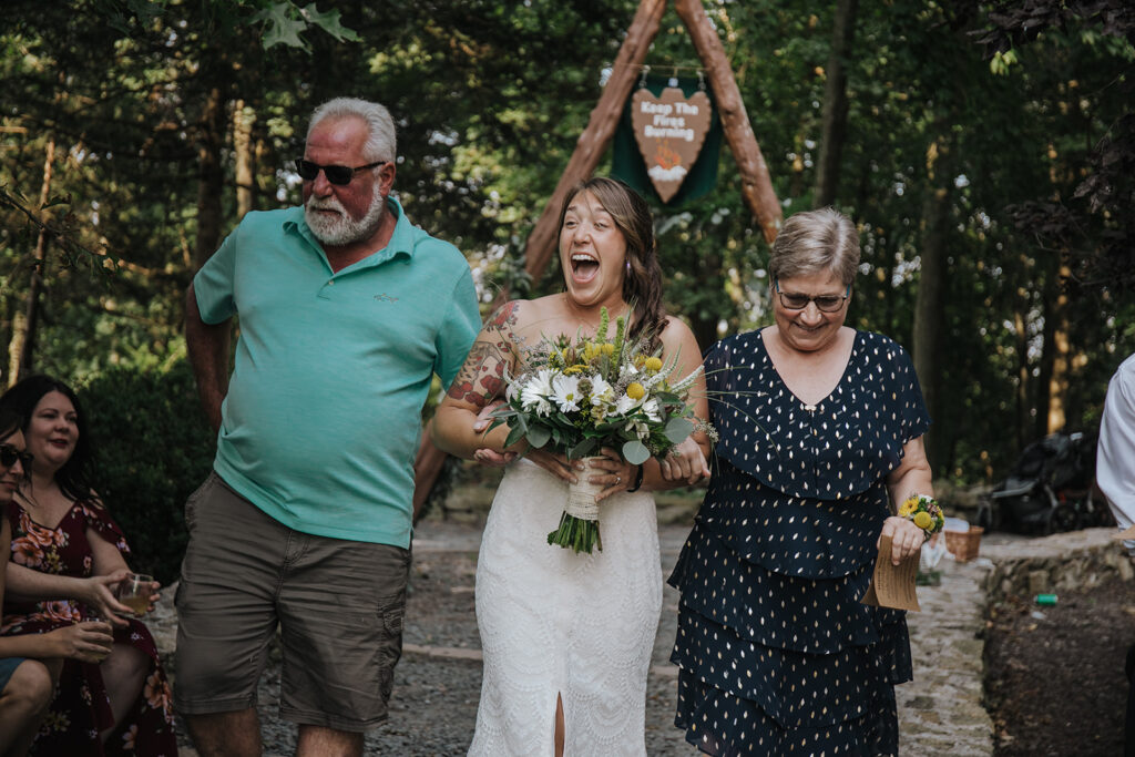 A bride and her parents walk down the aisle at her wedding. The bride is laughing, and her dad is wearing a green shirt. Her mom is wearing black with white polka dots. They are at a summer camp