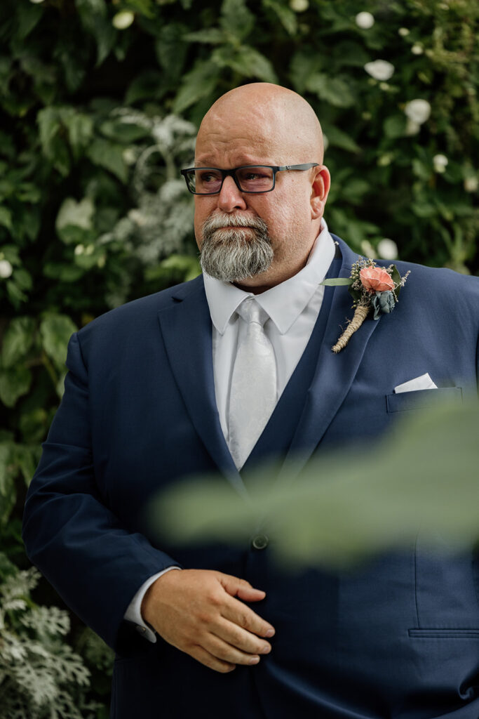 A groom is starting to tear up as his bride walks down the aisle. He is surrounded by greenery. He is wearing a navy suit.